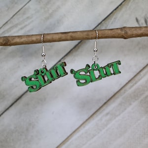May include: A pair of green wooden earrings spelling out "Slut" in a stylized font. The earrings are suspended from silver-colored fish hook ear wires and are displayed on a small wooden branch against a light background.