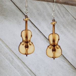 Puede incluir: Un par de pendientes de madera con forma de violonchelos. Los pendientes son de color marrón claro y tienen un acabado de madera natural.