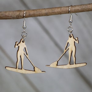 May include: A pair of wooden earrings featuring a silhouette of a person paddleboarding. The earrings are made of light brown wood and have silver hooks.