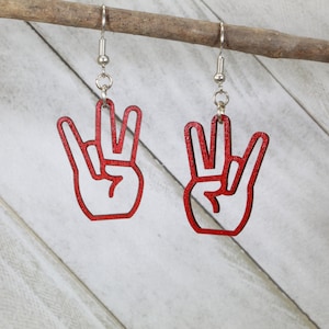 May include: A pair of red wooden earrings shaped like a hand giving the peace sign. The earrings are hanging from silver hooks.