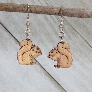 May include: A pair of wooden earrings featuring a squirrel design. The earrings are brown and have silver hooks.