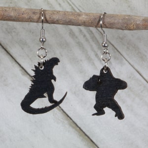 May include: A pair of black wooden earrings featuring a silhouette of Godzilla and King Kong. The earrings are attached to silver hooks.