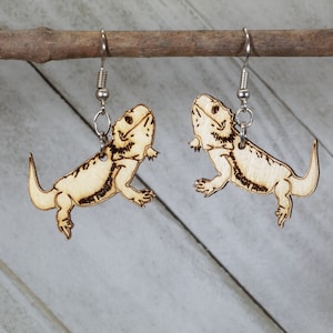 May include: A pair of wooden earrings featuring a bearded dragon design. The earrings are light brown and have silver hooks.