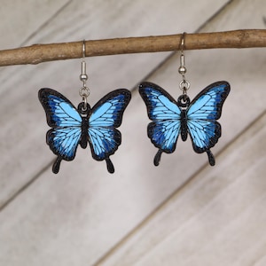 May include: A pair of butterfly earrings with a light blue and black design. The butterfly wings have a detailed pattern, and the earrings are suspended from silver-colored hooks. The earrings are displayed on a wooden branch.