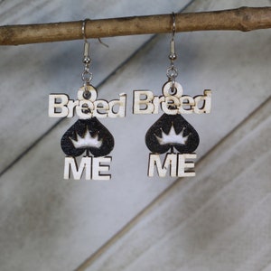 May include: Wooden dangle earrings with the words "Breed Me" in a playful font. The earrings feature a black spade shape with a white crown design. The earrings hang from silver-colored fish hook ear wires.