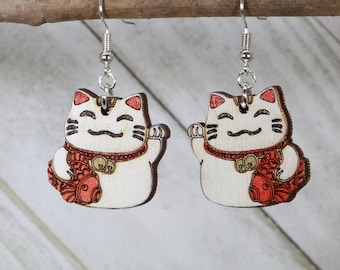以下が含まれることがあります： 赤い金色のディテールが施された白い木製の猫のチャームが付いた、銀色のドロップイヤリングのペア。猫は魚を持っており、首にベルが付いています。