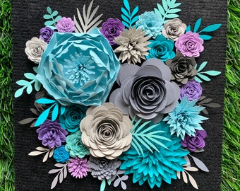 May include: A square black textured board adorned with a dense arrangement of layered paper flowers and foliage in shades of light blue, teal, dark gray, light gray, and purple. The composition features various petal shapes and sizes.