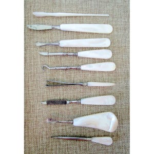 May include: A collection of nine manicure tools arranged vertically, each featuring a smooth, iridescent white mother-of-pearl handle. The set includes various metal implements such as a cuticle pusher, tweezers, a small brush, and a nail file.