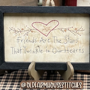 May include: A framed sign with the text "Friends Are The Stars That Twinkle in Our Hearts." The artwork features a heart and star design with a black frame. The sign is displayed on a wooden stand.