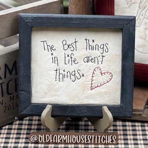 May include: A framed sign with the text "The Best things in Life aren't things" and a stitched red heart. The frame is black, and the sign is displayed on a wooden stand. The sign is on a checkered surface.