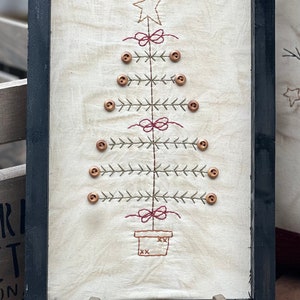 May include: A framed embroidery of a Christmas tree with a star on top, red bows, and brown buttons. The tree is in a pot with an embroidered design. The frame is black and the embroidery is on a white background. The image includes the text "@OLDFARMHOUSESTITCHES".