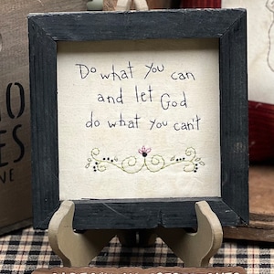 May include: A framed artwork with the text "Do what you can and let God do what you can't." The black framed art has a beige background with embroidered floral accents. The artwork is displayed on a wooden easel. The shop's name, @OLDFARMHOUSESTITCHES, is at the bottom.