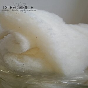 May include: A close-up of a large, white, fluffy pile of organic cotton filling. The cotton is packaged in a clear plastic bag. The text "I SLEEP SIMPLE Natural Organic Made in the USA" is visible at the top of the image.