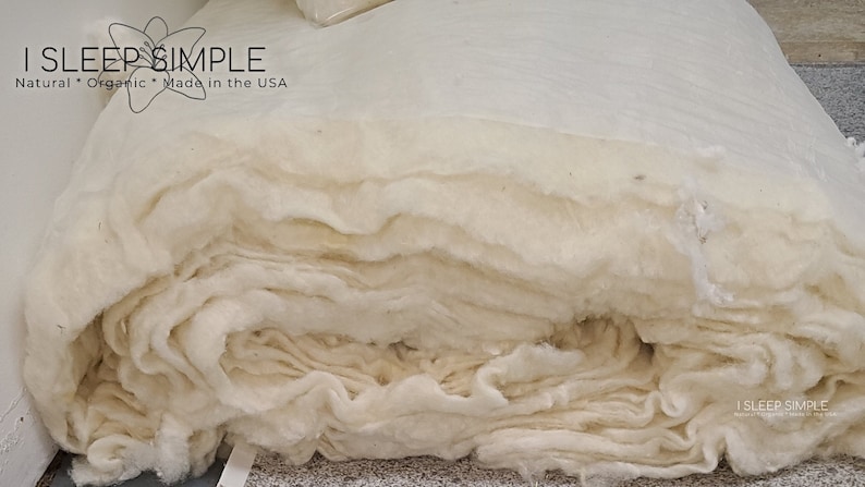 May include: A close-up of a large roll of white cotton batting. The batting is folded and stacked, creating a soft and fluffy texture. The label "I SLEEP SIMPLE Natural * Organic * Made in the USA" is visible on the top of the roll.