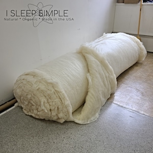 May include: A large roll of white cotton batting, unfurled and partially rolled up. The cotton is soft and fluffy, and the roll is sitting on a tiled floor. The text "I SLEEP SIMPLE Natural * Organic * Made in the USA" is visible in the background.