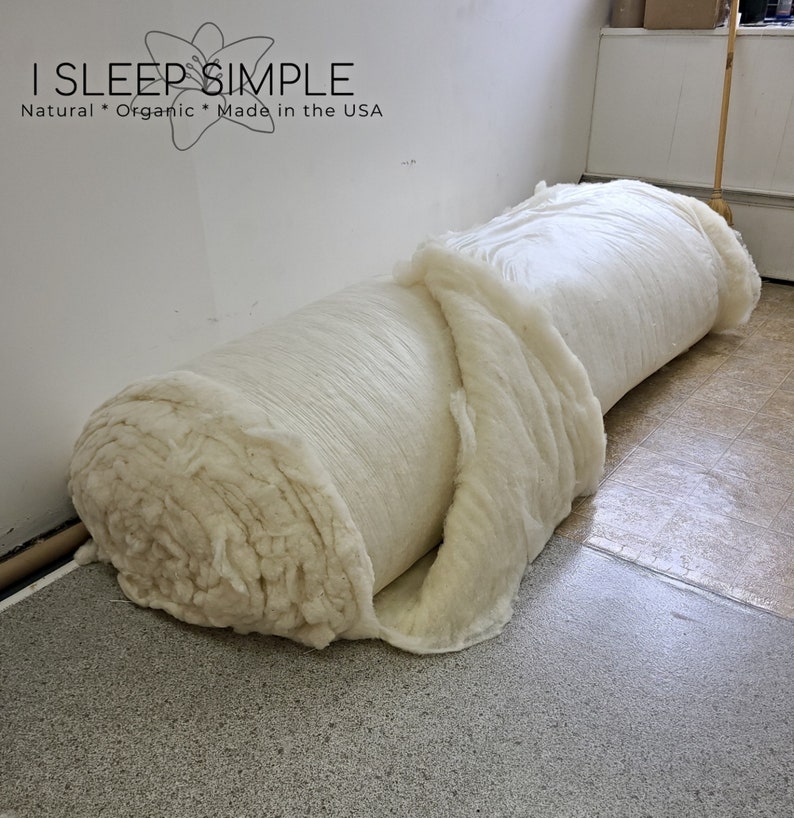 May include: A large roll of white cotton batting, unfurled and partially rolled up. The cotton is soft and fluffy, and the roll is sitting on a tiled floor. The text "I SLEEP SIMPLE Natural * Organic * Made in the USA" is visible in the background.