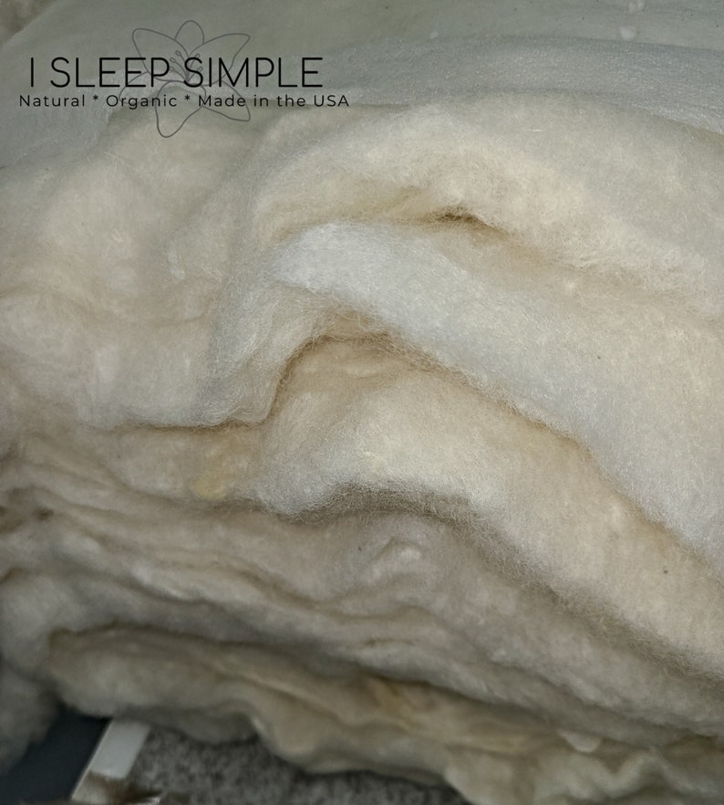 May include: A close-up of a stack of white, fluffy, natural wool batting. The wool is soft and airy, and it is perfect for stuffing pillows, cushions, and other home decor items. The label on the top of the stack reads "I Sleep Simple Natural * Organic * Made in the USA".
