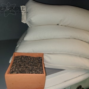 Peut inclure: Une pile de trois oreillers blancs en coque de sarrasin avec un pot en terre cuite brun rempli de coques de sarrasin devant eux. Les oreillers portent des &eacute;tiquettes indiquant "Buckwheat Hulls 100%".