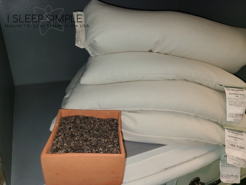 Peut inclure: Une pile de trois oreillers blancs en coque de sarrasin avec un pot en terre cuite brun rempli de coques de sarrasin devant eux. Les oreillers portent des &eacute;tiquettes indiquant "Buckwheat Hulls 100%".