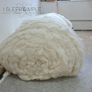 May include: A large, white, fluffy roll of organic cotton. The cotton is tightly packed and has a natural, unbleached appearance. The text "I SLEEP SIMPLE Natural * Organic * Made in the USA" is visible at the top of the image.