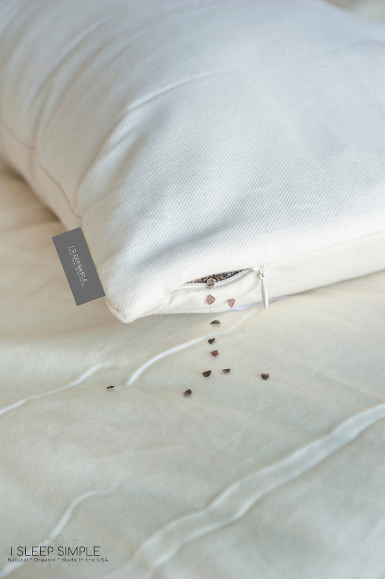 Peut inclure: Un oreiller blanc avec une fermeture &eacute;clair ouverte, laissant &eacute;chapper des coques de sarrasin sur un drap blanc. L'oreiller porte une &eacute;tiquette indiquant "I SLEEP SIMPLE - Natural Organic - Made in the USA".