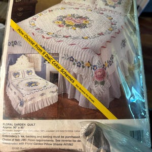 May include: A Bucilla stamped cross-stitch kit for a Floral Garden Quilt, measuring approximately 90 inches by 90 inches. The packaging displays a white quilt with floral embroidery, including pink and yellow roses, and matching pillow shams.