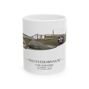 May include: A white ceramic mug featuring a side profile illustration of a P-40E Warhawk fighter plane with shark teeth nose art. Below the aircraft, the text reads "OLD EXTERMINATOR" P-40E WARHAWK COL. ROBERT L. SCOTT 23RD FIGHTER GROUP.