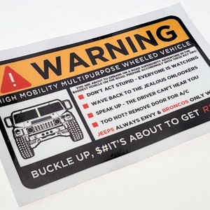 Puede incluir: Una pegatina rectangular con borde negro y sección superior amarilla que dice "WARNING". La pegatina presenta un gráfico Hummer y texto que incluye "BUCKLE UP" y "HIGH MOBILITY MULTIPURPOSE WHEELED VEHICLE".
