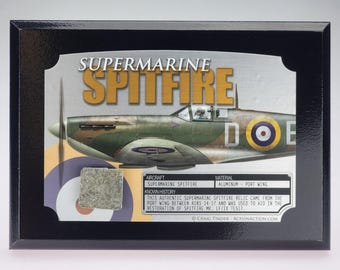 May include: A black wooden plaque featuring a Supermarine Spitfire aircraft illustration with a metallic relic piece attached. The text reads Supermarine Spitfire and includes a history box detailing the aluminum port wing relic from Spitfire Mk. LF/IX TE517.