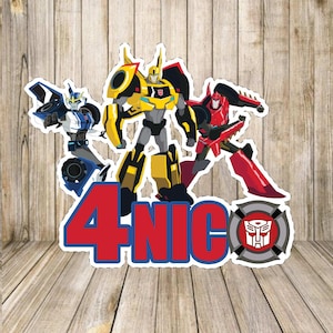 Pode incluir: Um topo de bolo de aniversário com três robôs Transformers, incluindo Bumblebee, em amarelo, azul e vermelho. O topo também inclui o número "4" e o nome "NIC" em vermelho com contornos azuis.