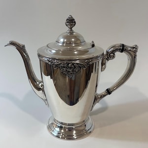 May include: A silver-colored coffee pot with a decorative floral design and a curved spout. The pot has a lid with a finial and a curved handle with black accents. The pot is on a pedestal base.