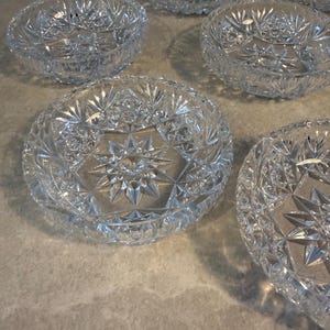 May include: A collection of clear, cut-glass bowls with starburst and geometric patterns. The bowls are round with a slightly flared rim. The cut glass reflects light, creating a sparkling effect. The bowls are arranged on a neutral-colored surface.