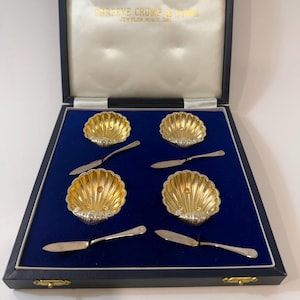 May include: A set of four gold-coloured shell-shaped dishes with silver trim, each with a small silver butter knife. The set is presented in a navy blue box with the text "SHREEVE CRUMP & LOWE JEWELER SINCE 1850".