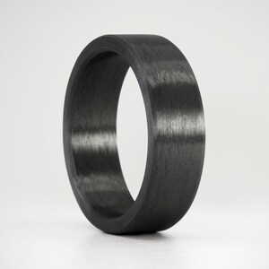 Puede incluir: Un anillo negro de fibra de carbono. El anillo tiene un acabado liso y mate y un perfil redondeado. El color gris oscuro es consistente en todo el anillo, con sutiles variaciones en la veta del material. El anillo es un diseño de banda simple.