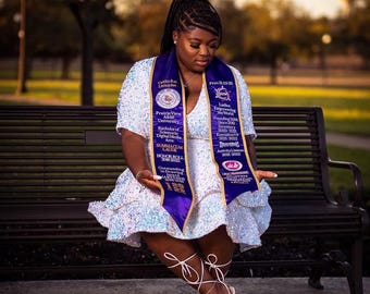 May include: A woman wearing a white sequined dress and a purple and white sash sits on a park bench. The sash has text and logos. She wears white strappy heels. The background is blurred with trees and a sunset.