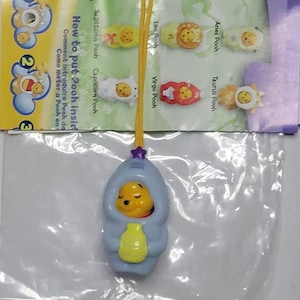 Può includere: Un ciondolo blu di Winnie the Pooh con una stella gialla in cima. Il ciondolo è attaccato a uno stringa gialla. Il ciondolo è in una busta di plastica con una carta che dice "Come mettere Pooh dentro".