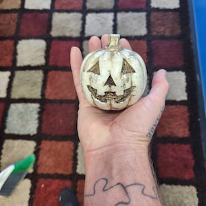 May include: A white ceramic pumpkin with a carved jack-o'-lantern face. The pumpkin is being held in a person's hand.
