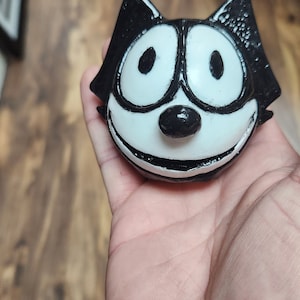 May include: A black and white ceramic figurine of a cartoon cat with a wide smile and large eyes. The figurine is held in a person's hand.