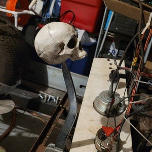 May include: A white skull shifter knob with a metal rod attached to a metal base. The shifter is mounted on a metal surface with wires and other mechanical parts visible.