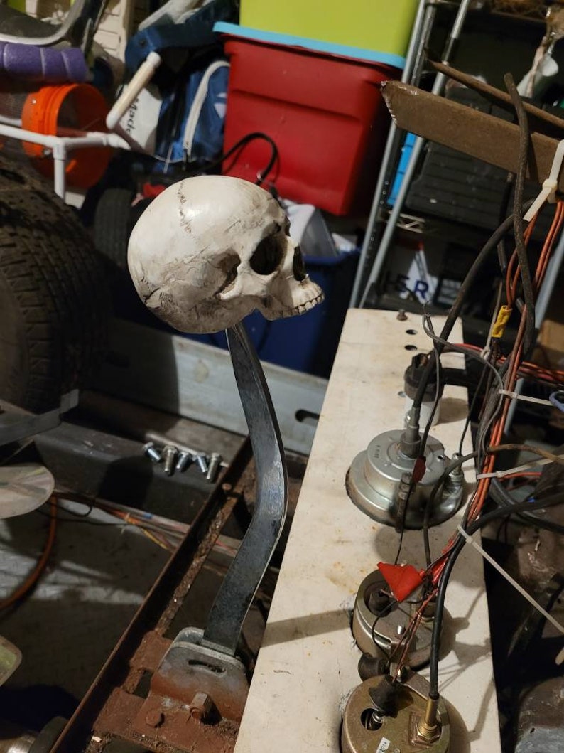 May include: A white skull gear knob with a metal rod attached to a metal base. The shifter is mounted on a metal surface with wires and other mechanical parts visible.