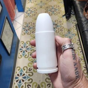 May include: A white, cylindrical object with a rounded top and a base, held in a hand. The object appears to be a container or applicator. The background includes a patterned rug and a tiled floor.