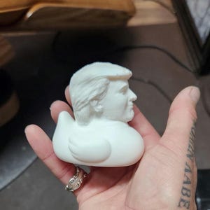 May include: White rubber duck with a detailed face of a man with blonde hair. The duck is being held in a hand with a tattoo.