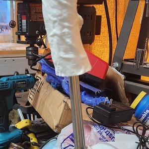 May include: A white, 3D printed,  gear shift knob with a textured surface. The knob is attached to a silver metal shaft and a black base. The base is surrounded by a variety of objects, including a blue and yellow drill, a white plastic bag, and a black power adapter.