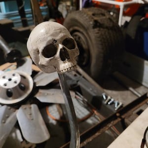 May include: A white skull gear knob for a car. The skull is detailed with a textured surface and is mounted on a silver metal rod.