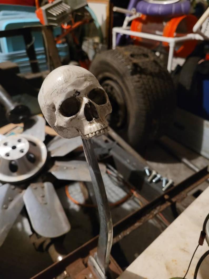 May include: A white skull gear knob for a car. The skull is detailed with a textured surface and is mounted on a silver metal rod.