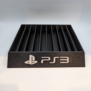 Puede incluir: Soporte de plástico negro con ranuras para guardar discos de juegos de video. El soporte tiene el logotipo de PlayStation y el texto "PS3" en la parte delantera.