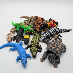 Puede incluir: Una colección de lagartos de juguete coloridos en varios patrones y colores, incluyendo verde, azul, negro, amarillo y naranja. Los reptiles están dispuestos sobre una superficie blanca.