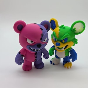 Könnte beinhalten: Zwei stilisierte Bärenfiguren. Einer ist rosa und lila, mit genähten Nähten und einem wütenden Gesichtsausdruck. Der andere ist grün, gelb und blau, ebenfalls mit einem wütenden Gesichtsausdruck. Beide haben übertriebene Merkmale und einen cartoonartigen Stil.