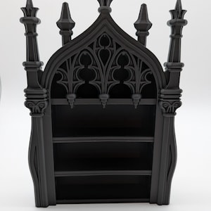 May include: A black, gothic-style bookshelf with three shelves. The top features ornate, arched detailing and decorative finials. The bookshelf is made of a solid material and stands on a white surface.