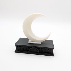 May include: A cream-colored crescent moon sculpture atop a black rectangular base with arched detailing. The sculpture is set against a white background, creating a minimalist aesthetic. The base has a decorative, gothic-inspired design.